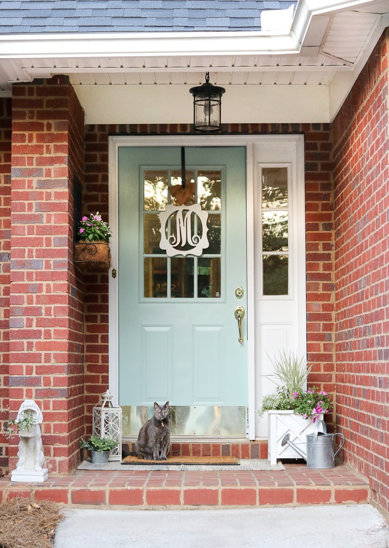 Small front porch decorating ideas with a layered rug, container garden of plants and flowers, watering can, statue of a dog holding flowers, and monogram wreath.