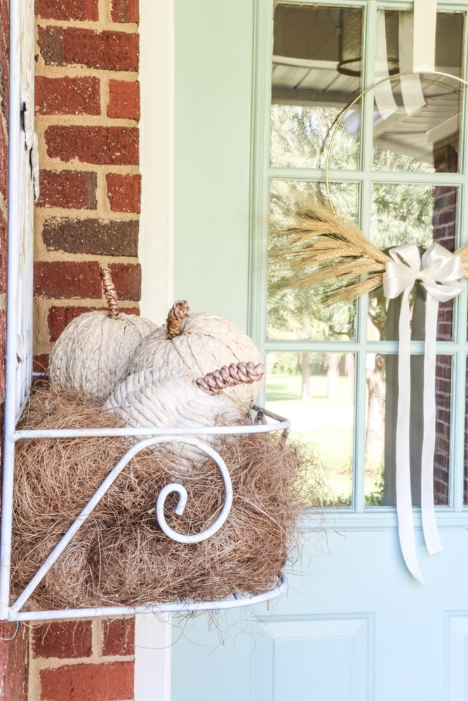 Faux pumpkins wrapped in a natural twine sitting inside a iron wall flower hanger on a front porch for fall.