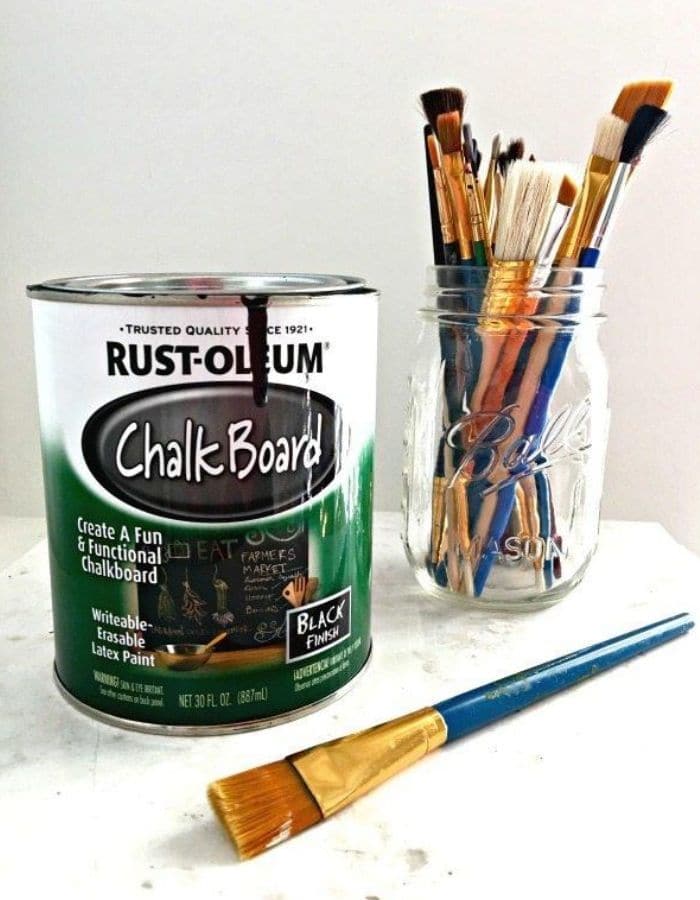 In this image there is a small can of Rust-Oleum Chalkboard Paint next to a mason jar full of artist paint brushes.