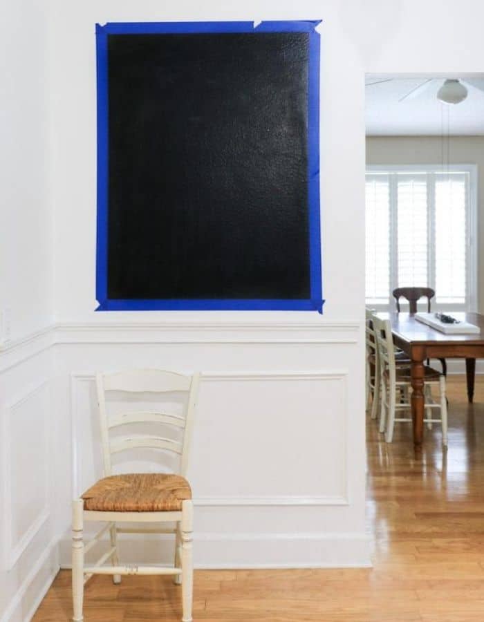 There is a small wall with a large rectangle of painters tape on the wall. Painted within the tape is a black chalkboard.
